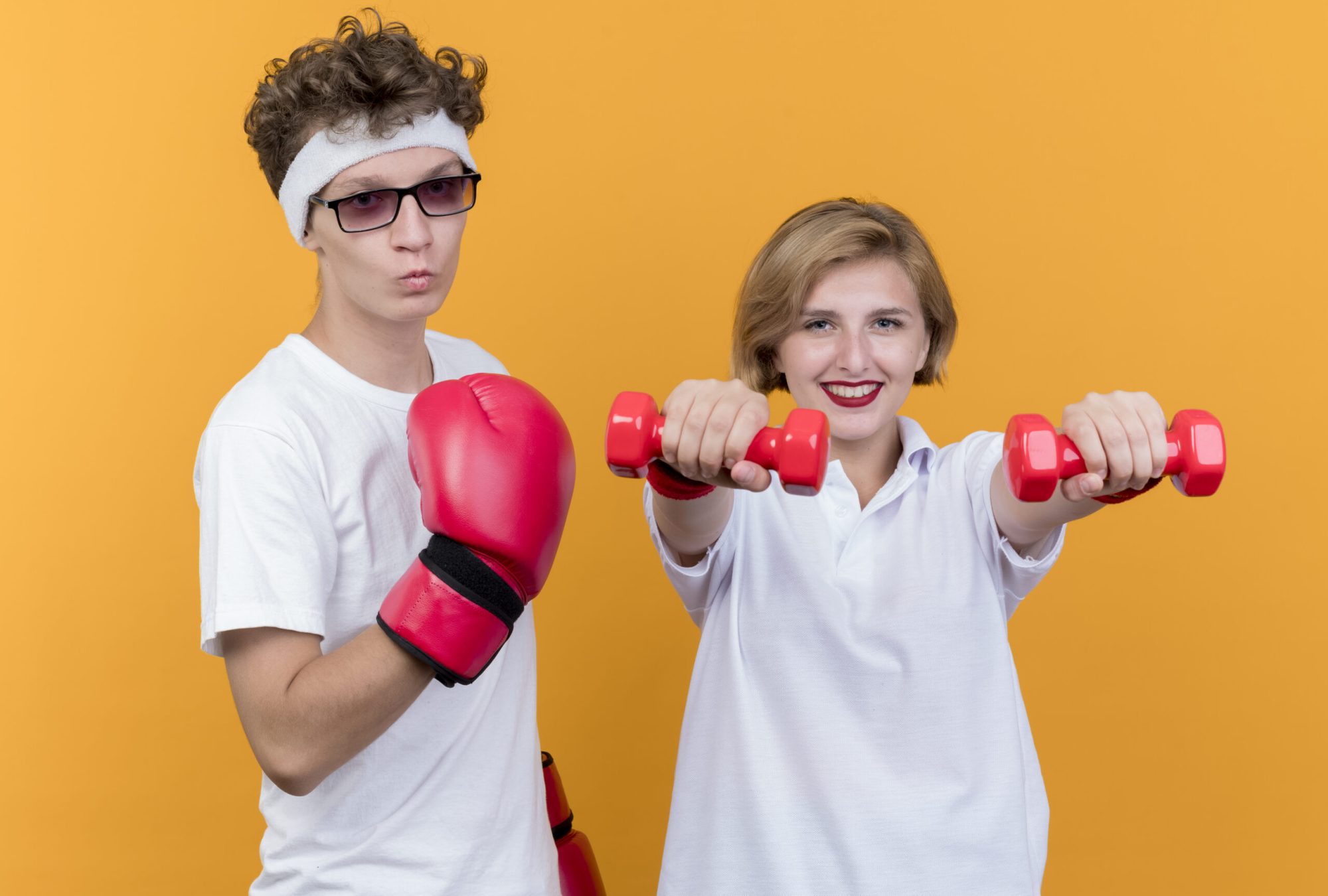 young sporty couple woman with dumbbells standing next to her boyfriend with boxing gloves posing looking at camera standing over orange background